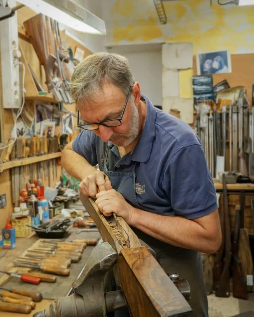 Armurier en lunettes sculptant une crosse de fusil en bois avec un ciseau dans son atelier. Outils visibles.