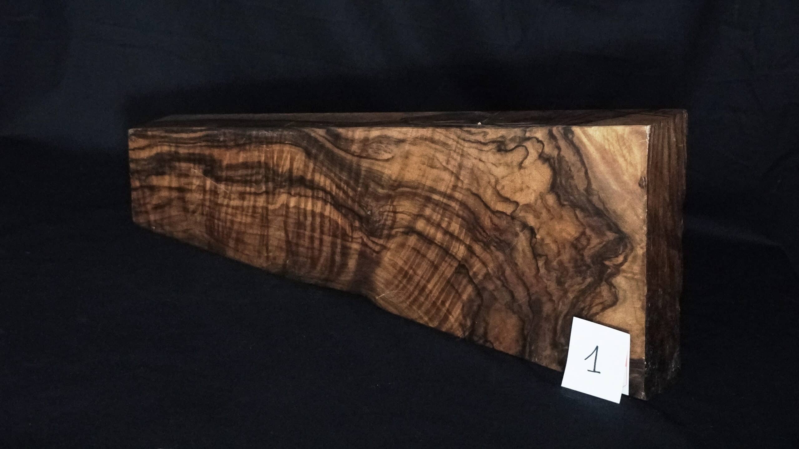 Bloc de bois brut foncé avec grain ondulé naturel et marques sombres distinctives, étiqueté '1' sur fond noir.