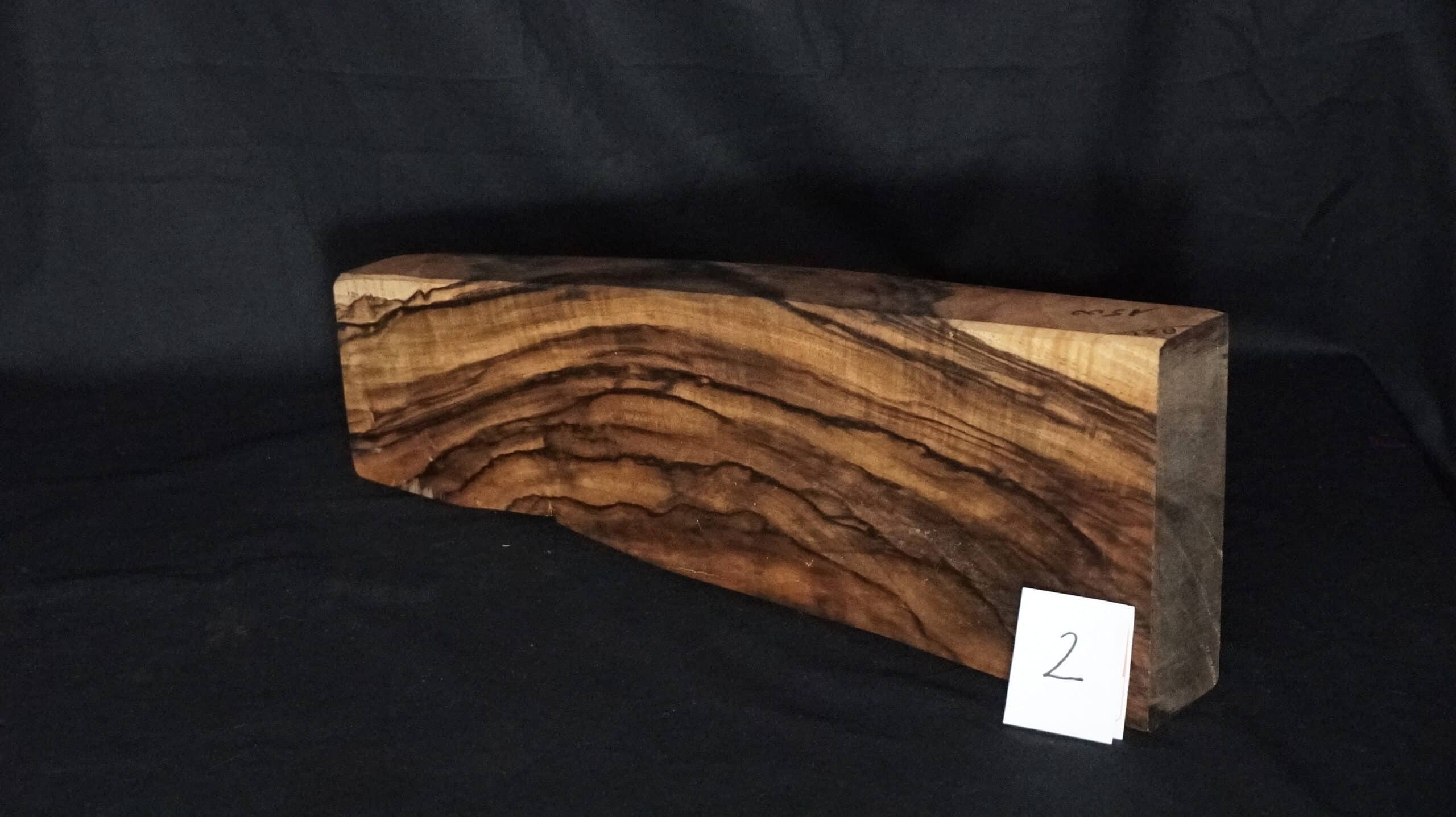 Bloc de bois d'olivier massif aux veines ondulées sombres sur fond noir. Étiquette blanche avec le numéro 2.