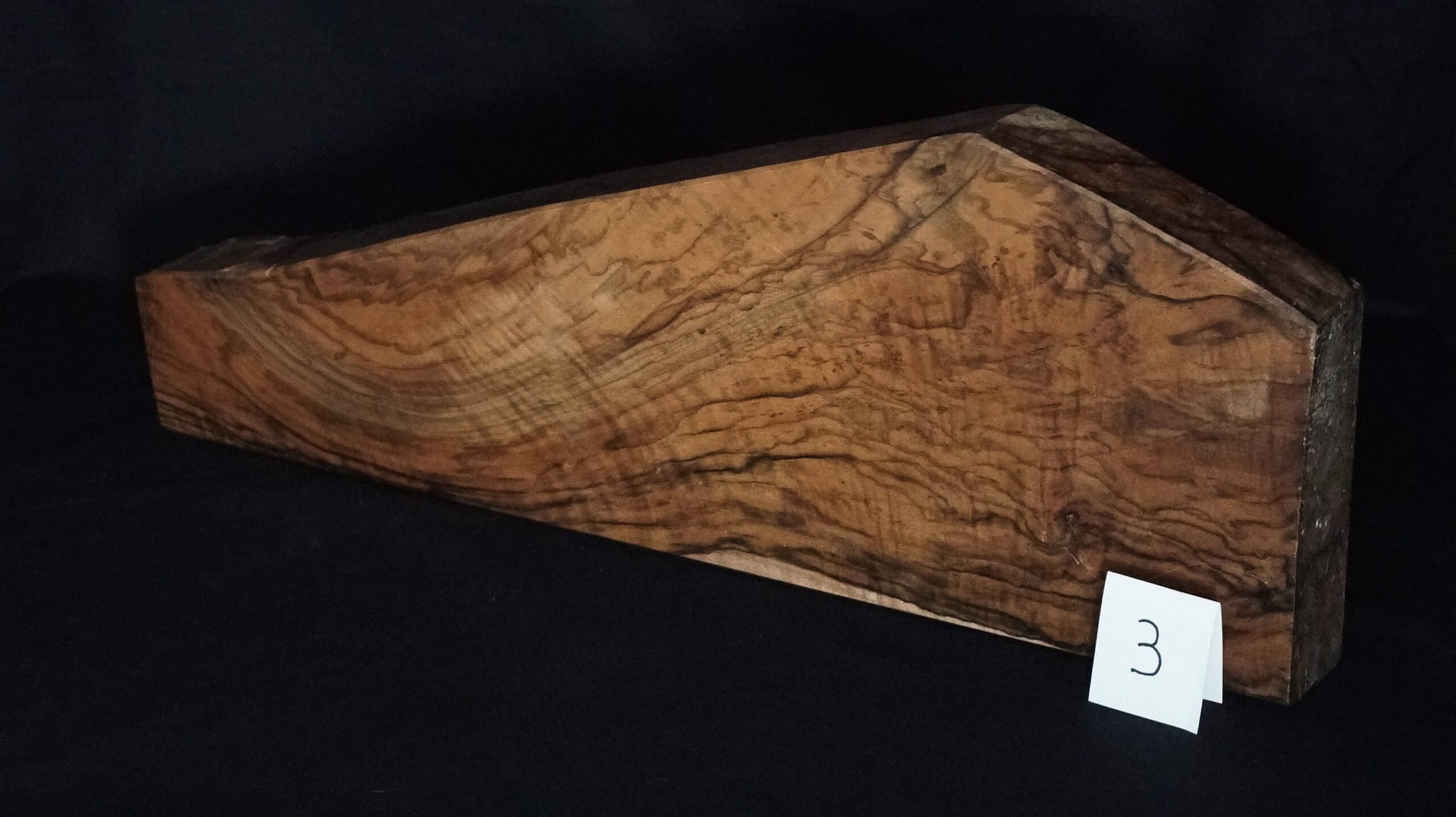 Bloc de bois brun de ronce ou de loupe, à la forme de toit et au grain ondulé prononcé, sur fond noir. Une étiquette "3" est visible.