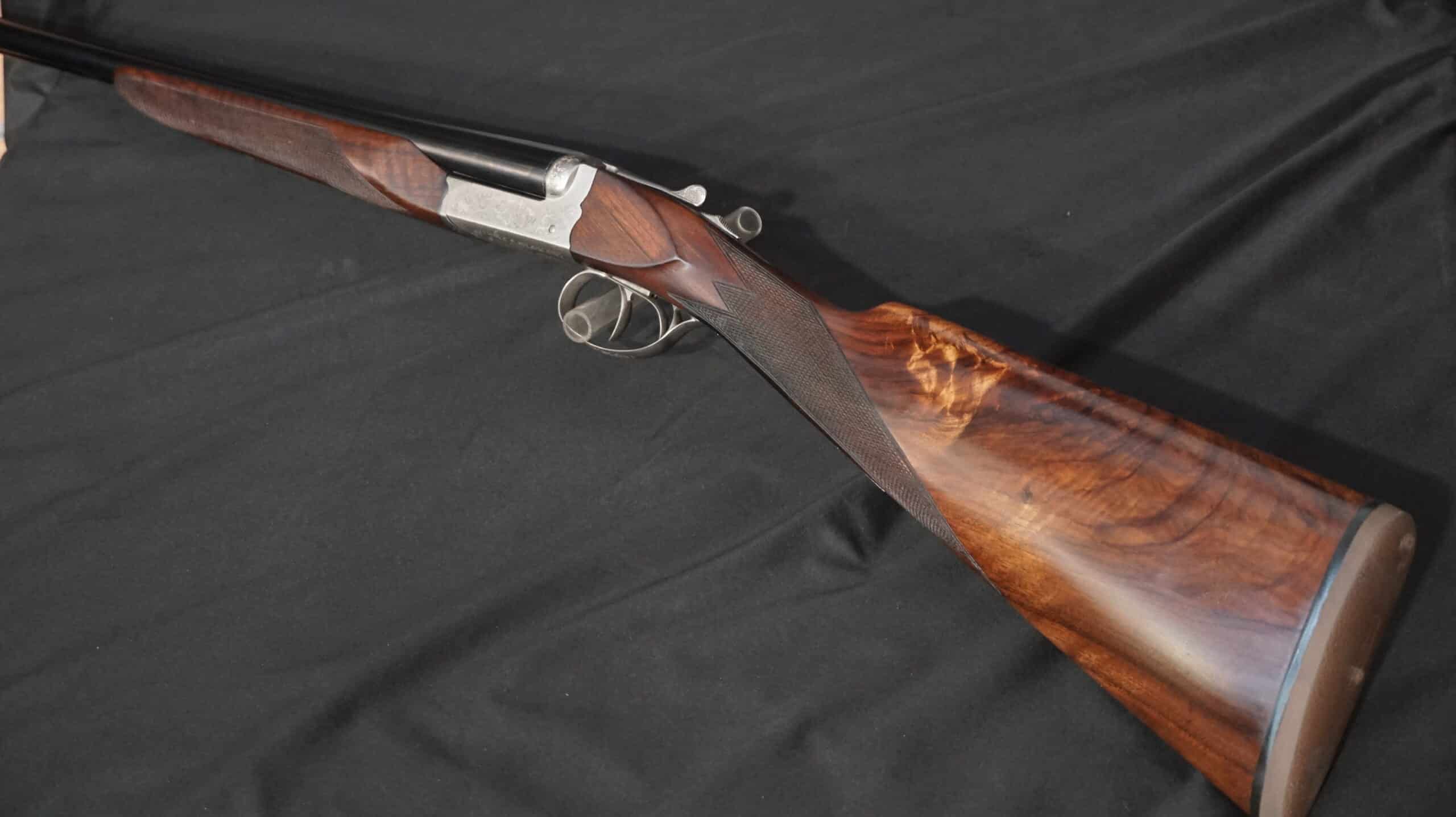 Fusil de chasse juxtaposé de luxe avec crosse en noyer veiné et bascule gravée sur fond noir.