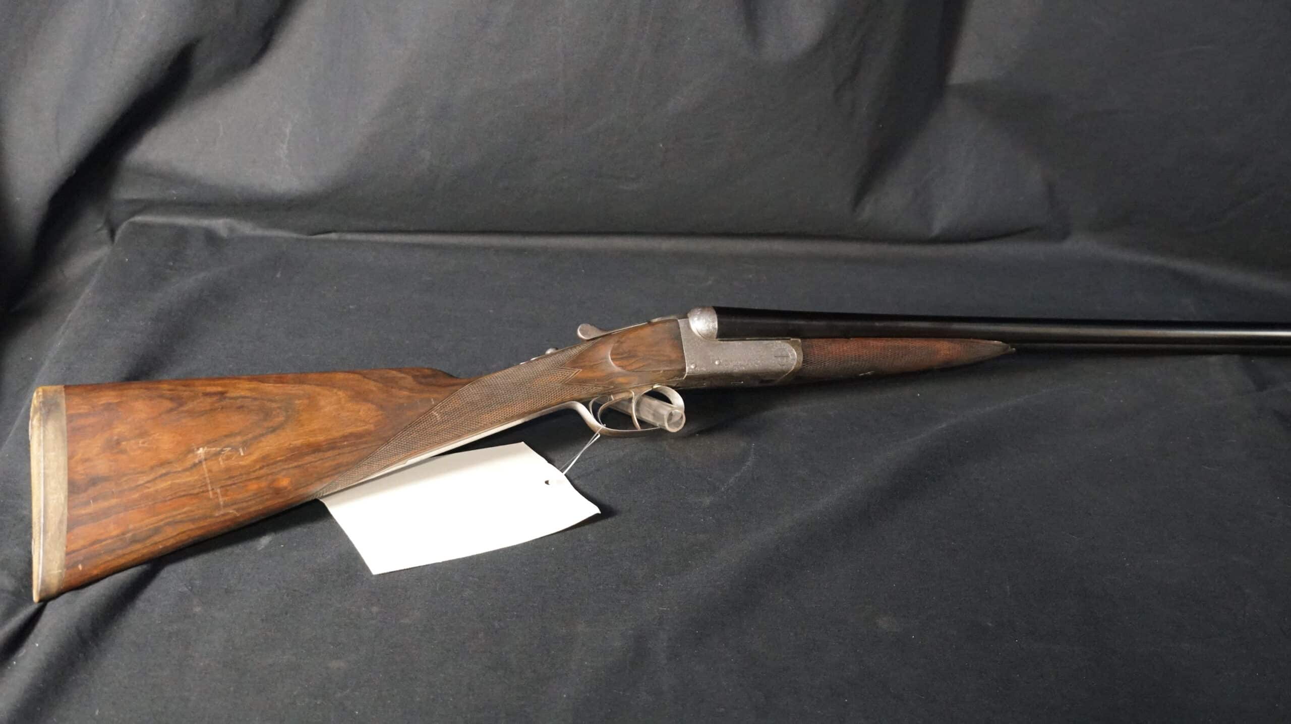 Fusil de chasse juxtaposé ancien, crosse en bois quadrillée, double détente, canon noir sur fond textile sombre.