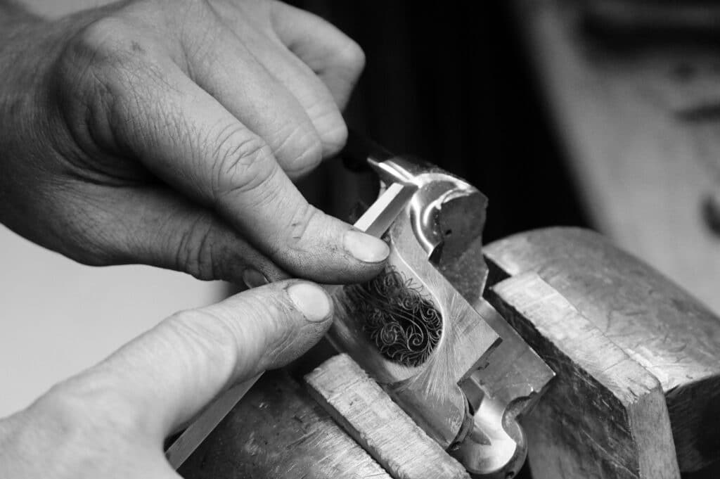 Mains d'artisan gravant un motif ornemental sur une pièce métallique tenue dans un étau. Travail de précision manuel.