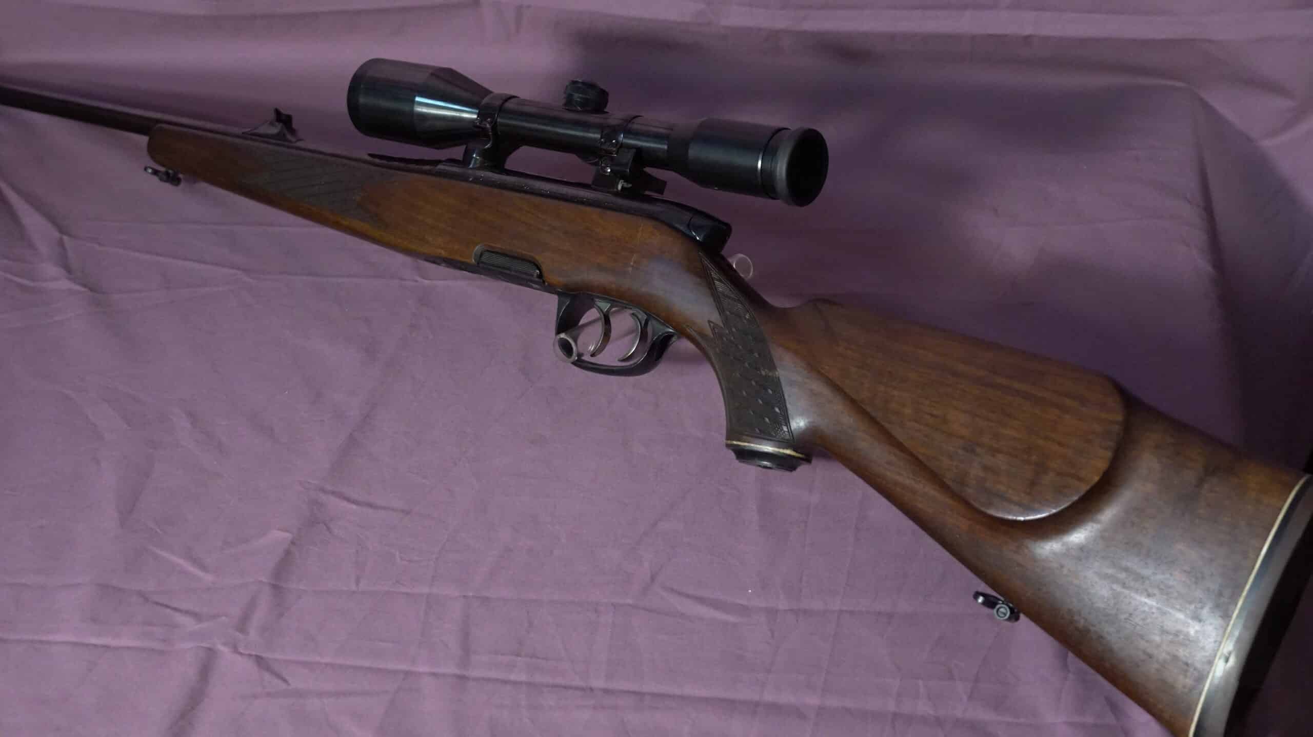 Carabine de chasse en bois foncé avec lunette de visée noire et détente, posée sur un fond de tissu mauve froissé.