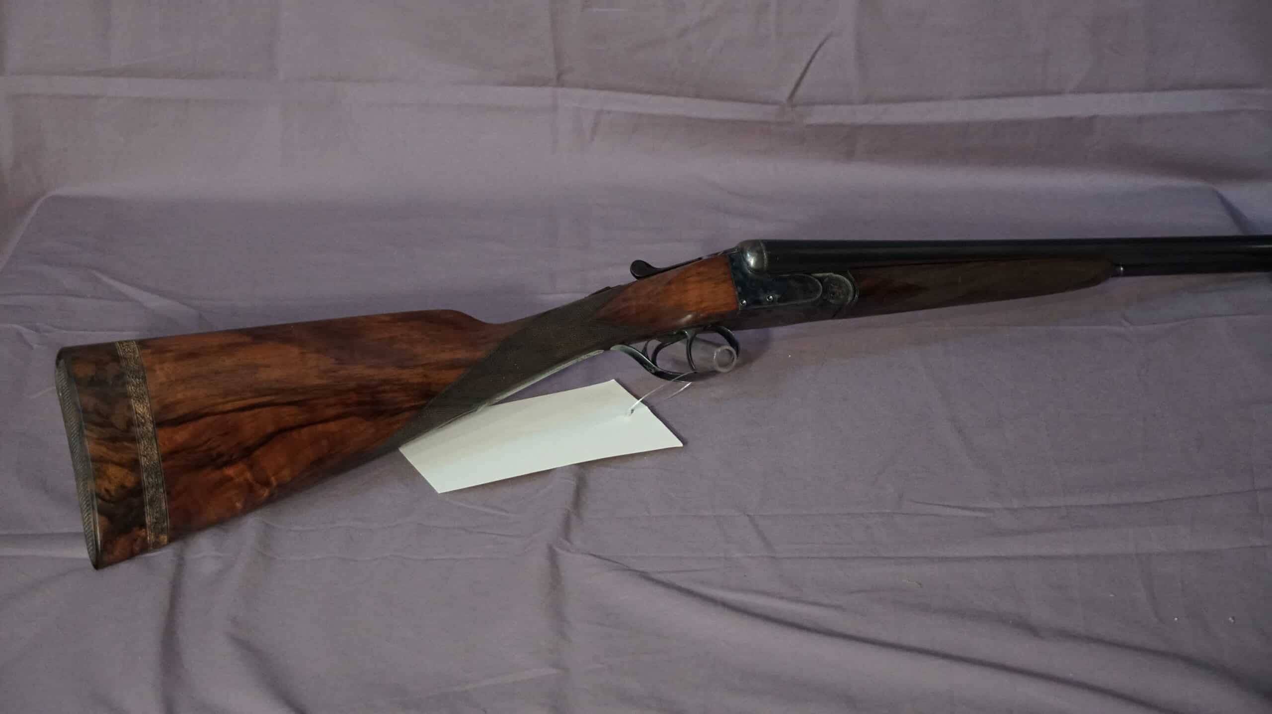 Canon de fusil juxtaposé avec crosse en bois foncé richement veiné et plaque de couche décorée, posé sur un drapé mauve.