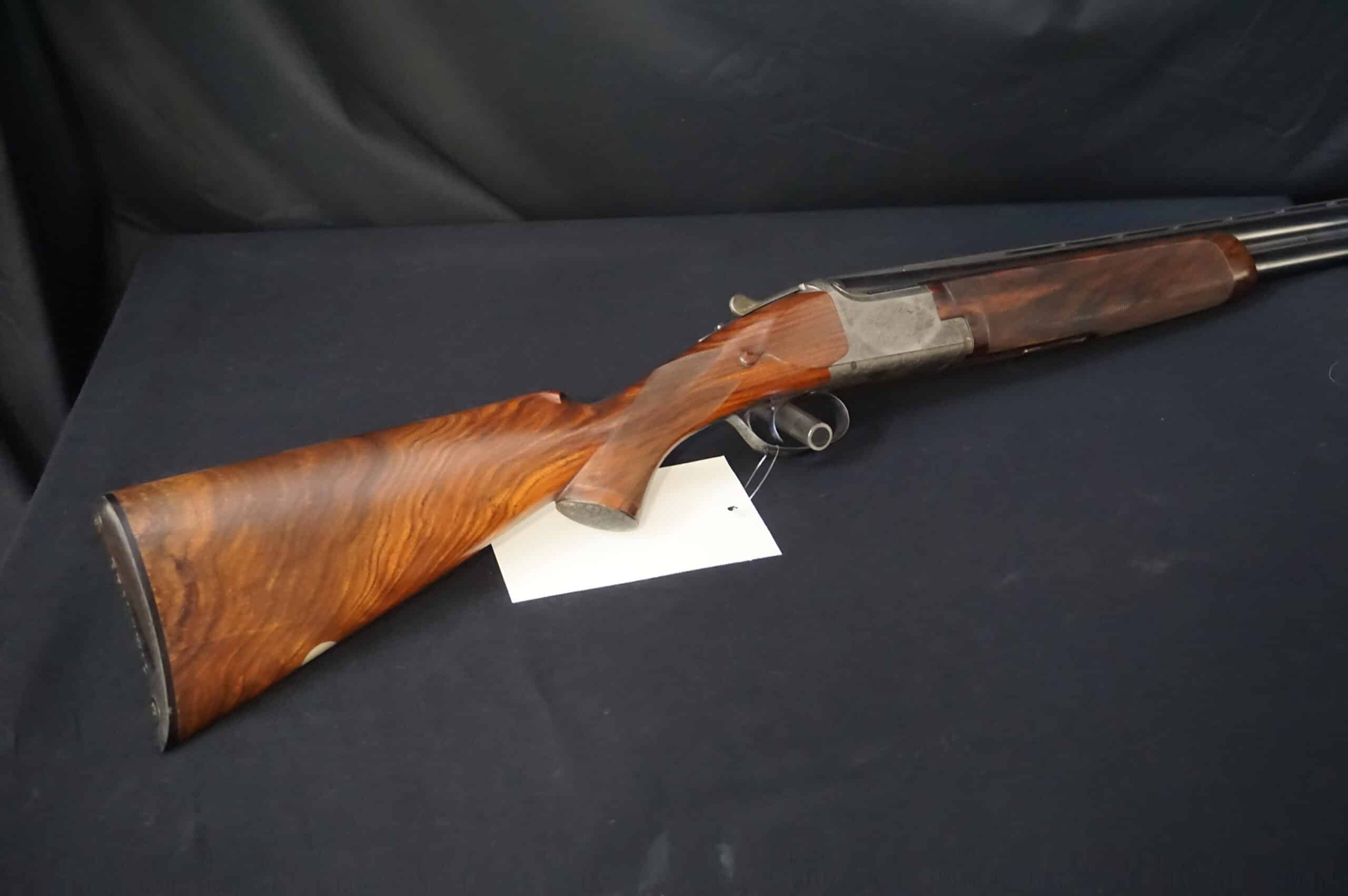 Fusil superposé, crosse en noyer aux veines prononcées, contre un fond sombre. Une étiquette blanche est attachée.