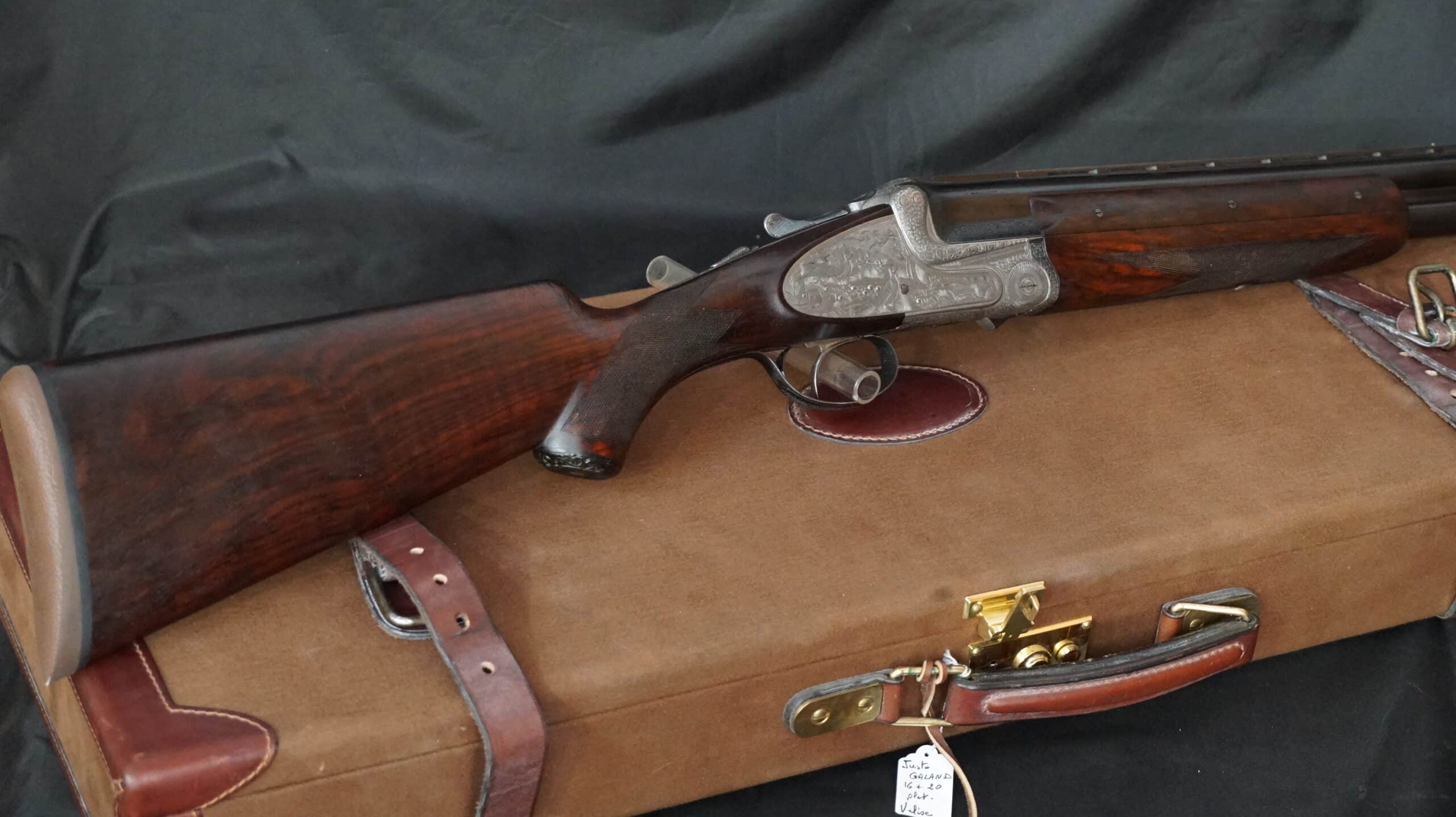 Fusil juxtaposé gravé (Justo Galand 16+20) sur une mallette marron avec poignées et boucles en cuir. Étiquette visible.