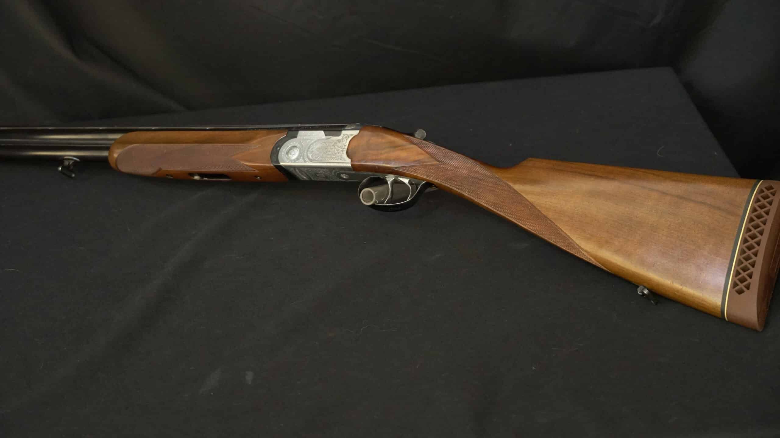 Fusil juxtaposé avec bois de noyer et gravures fines sur la bascule, contre un fond noir.