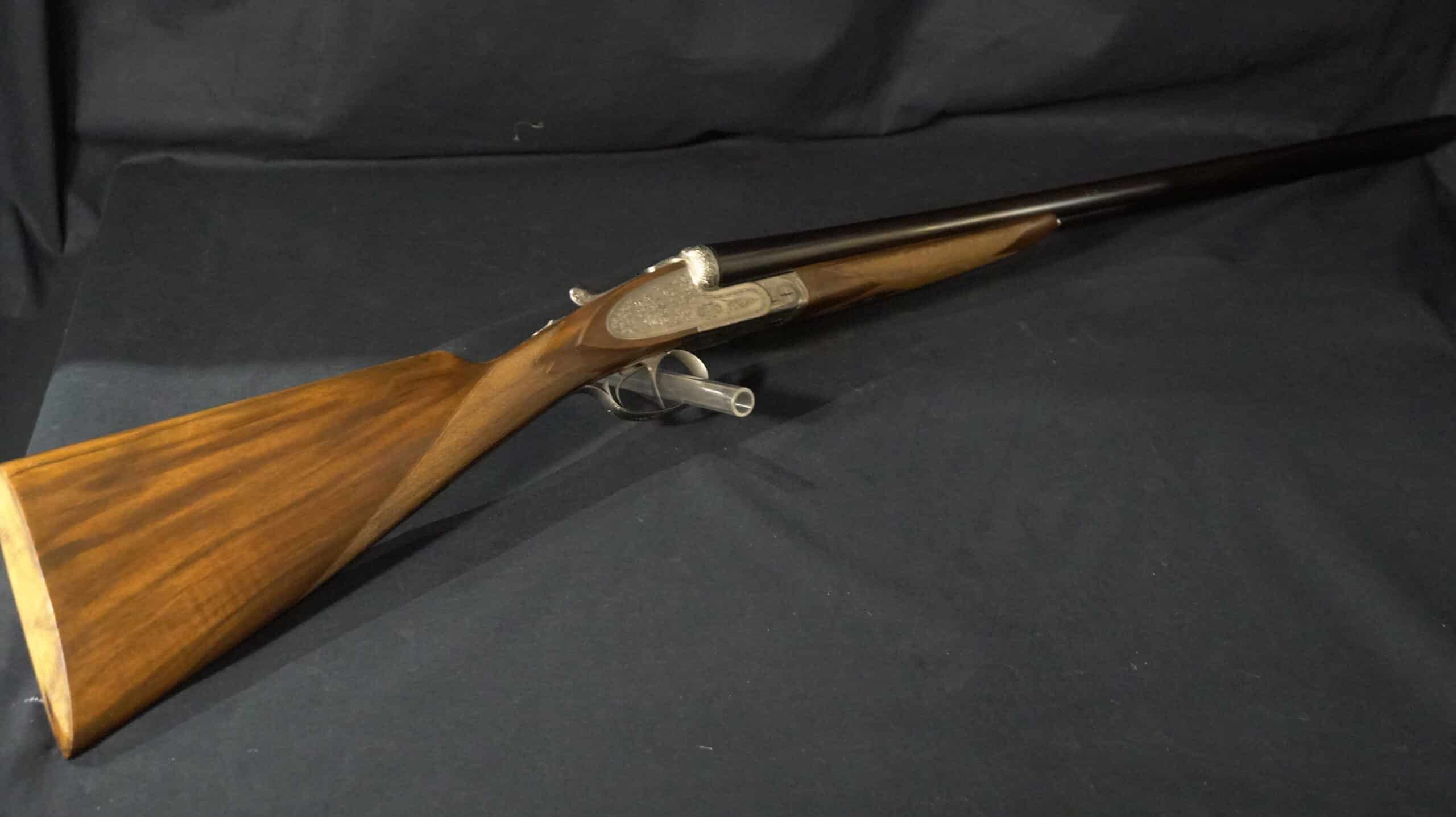 Fusil juxtaposé classique à platine gravée, crosse en noyer richement veiné, reposant sur un fond noir uni.
