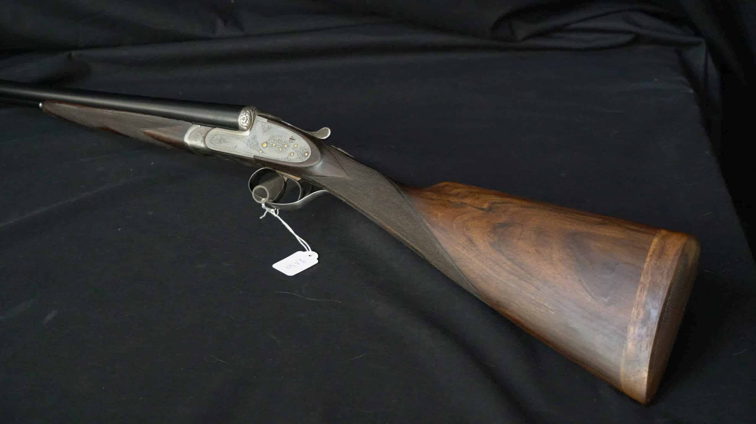 Fusil de chasse juxtaposé avec gravures dorées sur la bascule et crosse en bois précieux sur fond noir.