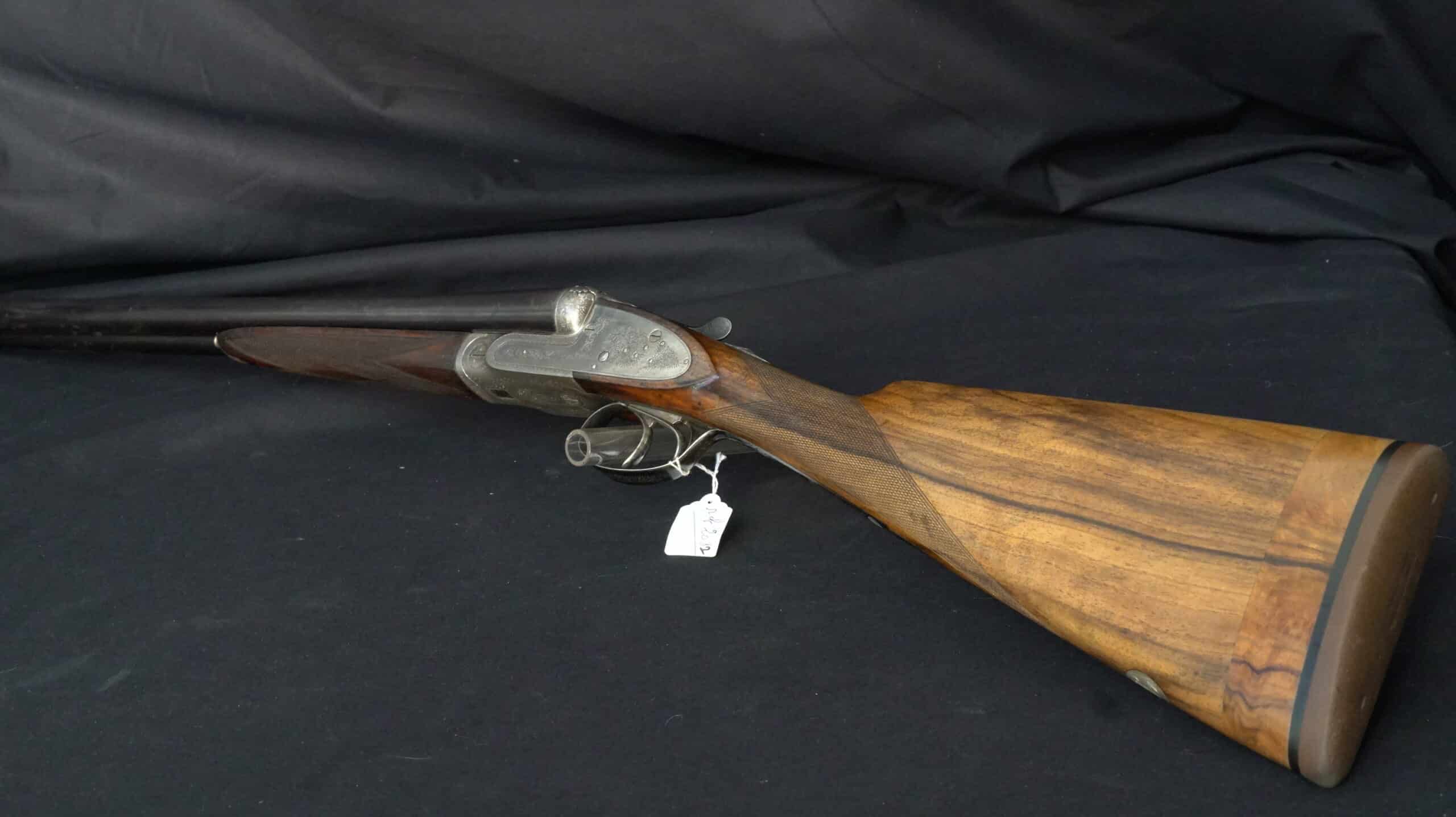 Fusil de chasse juxtaposé ancien avec crosse en bois de noyer et bascule ciselée sur fond textile noir.