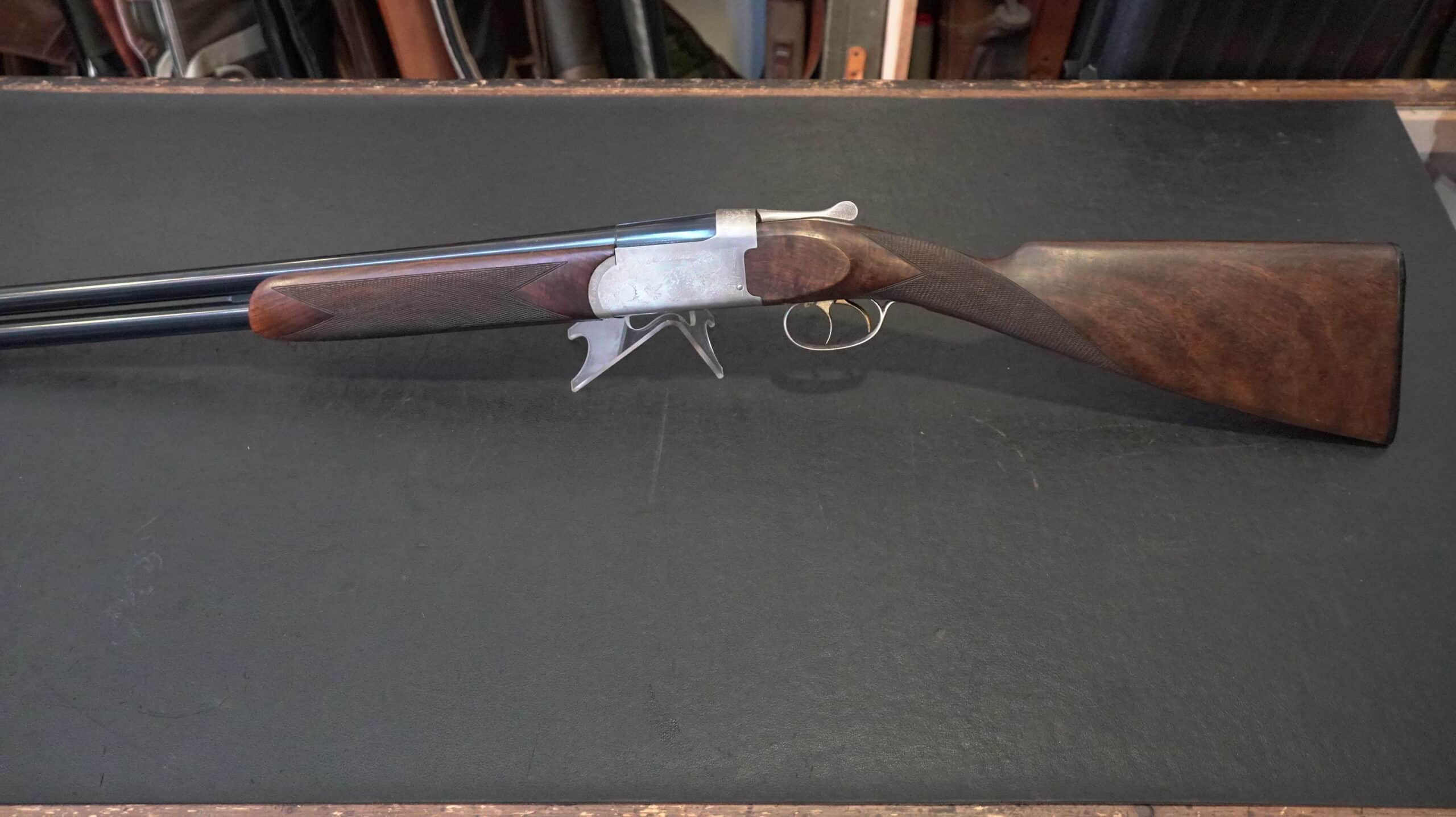Fusil de chasse superposé avec crosse en bois de noyer et bascule argentée gravée sur fond noir.