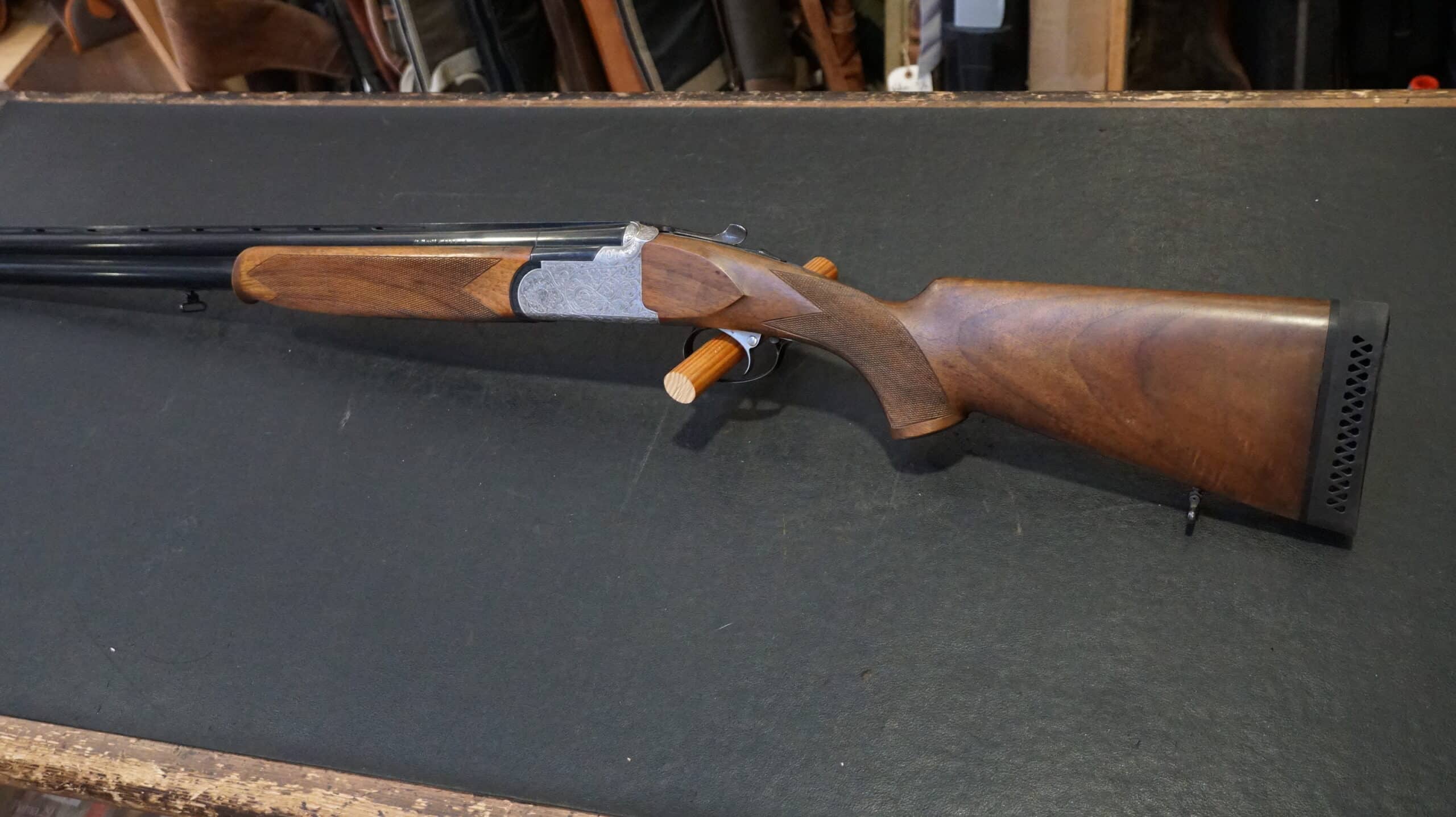Fusil de chasse superposé avec crosse en bois et bascule gravée, posé sur un comptoir noir.